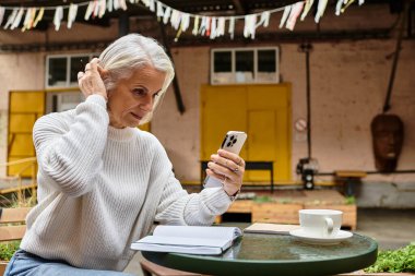 Şık, olgun bir kadın akıllı telefonuyla meşgul olurken kahvenin ve notların tadını çıkarıyor..