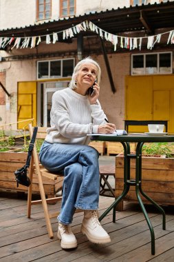 A beautiful older woman with gray hair sits at a table, engaged in a phone conversation. clipart