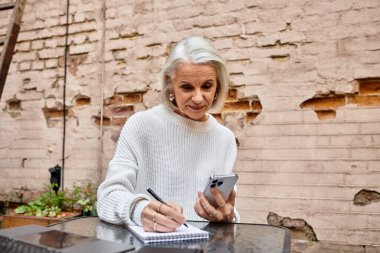 Gri saçlı, güzel, yaşlı bir kadın telefonuna bakarken düşüncelerini not alıyor..
