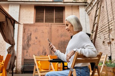 Güzel gri saçlı olgun bir kadın bir kafede rahatça oturup, akıllı telefonuyla meşgul olur..