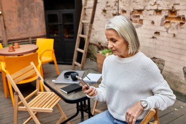 Büyüleyici bir açık hava ortamında, gri saçlı bir kadın akıllı telefonunu kullanırken rahatlar..