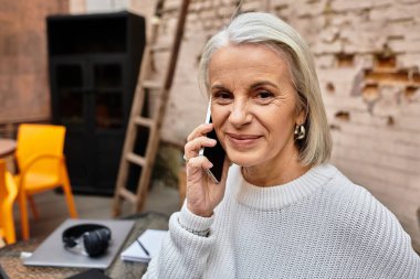 Samimi bir kafede, olgun bir kadın gülümseyerek hoş bir telefon görüşmesi yapar..