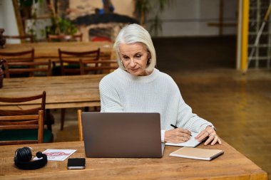 A mature woman writes notes while focused on her laptop in a warm cafe atmosphere. clipart