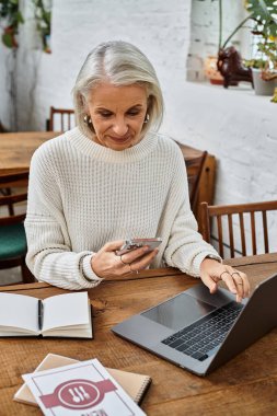 Güzel, gri saçlı bir kadın, kırsal bir kafede akıllı telefonu ve laptopuyla birden fazla iş yapıyor..