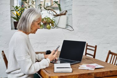 Gri saçlı, güzel, olgun bir kadın rahat bir iş yerinde akıllı telefonuyla etkileşime giriyor..