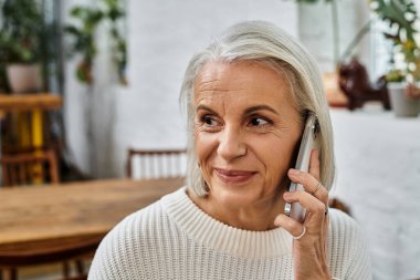 A mature woman with beautiful gray hair enjoys a lively conversation on her phone while smiling. clipart