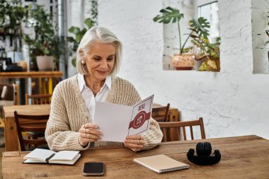 A lovely gray haired woman sits at a wooden table, absorbed in the cafe menu under soft lighting. clipart