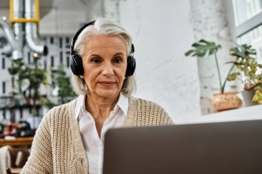 A mature woman with gray hair listens attentively during her virtual gathering in a warm atmosphere. clipart