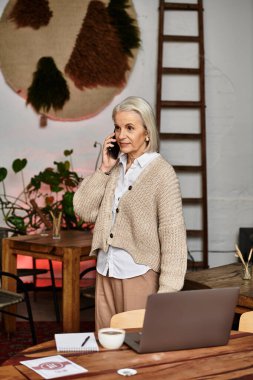 A beautiful mature woman talks on the phone while standing in a warm, inviting workspace. clipart