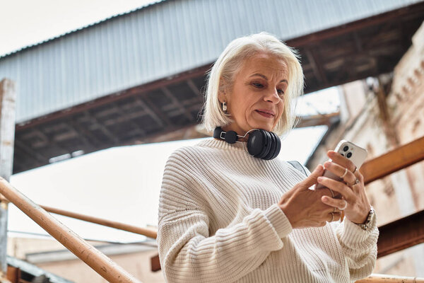 A beautiful older woman with gray hair relaxes on wooden steps as she texts on her phone.