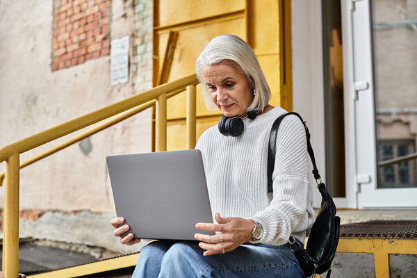 A mature woman with gray hair is engaged in her laptop work while resting on a bright staircase.