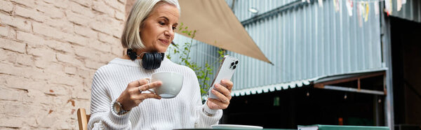 A beautiful mature woman with gray hair drinks coffee and checks her smartphone at a cafe.