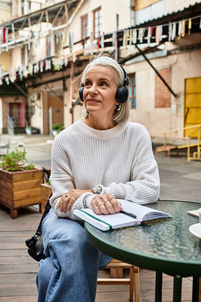 Красивая зрелая женщина носит наушники и пишет в блокноте, расслабляясь на открытом воздухе.