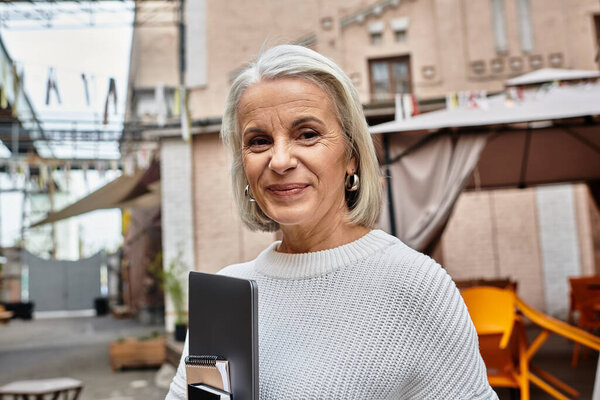A charming mature woman with gray hair stands in an urban courtyard, exuding warmth and confidence.