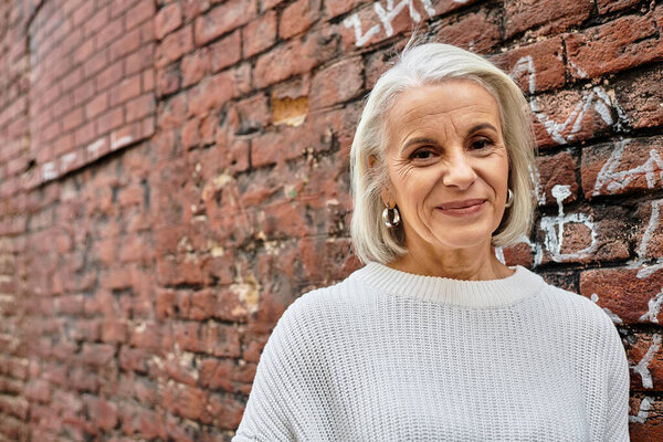 A charming older woman with silver hair leans against a brick wall, enjoying the sunlight.