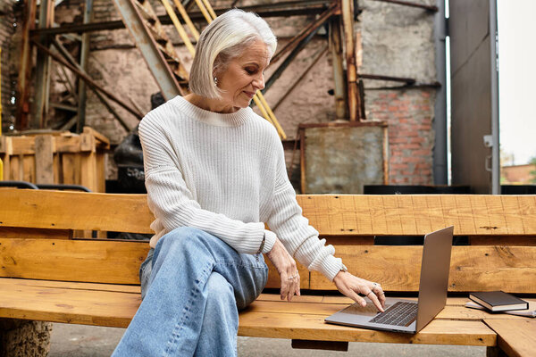 A beautiful mature woman with gray hair utilizes her laptop while sitting on a wooden bench.