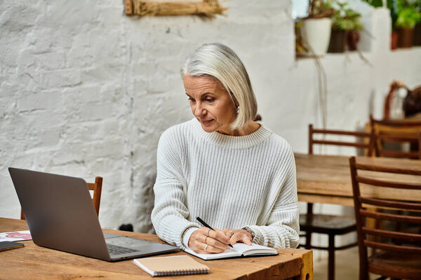 Взрослая женщина с седыми волосами записывает заметки во время общения с ноутбуком в теплом кафе.