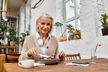 A smiling woman with gray hair savors her food in a bright, plant filled cafe. clipart