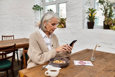 Güzel, olgun bir kadın bir kafede yemeğinin tadını çıkarırken akıllı telefonuyla meşgul olur..