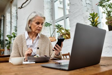 Güzel gri saçlı olgun bir kadın yemeğinin tadını çıkarıyor ve rahat bir masada akıllı telefonunu kontrol ediyor..
