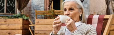 The mature woman savors her hot beverage in a cozy garden nook under soft lighting. clipart