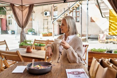 Gri saçlı olgun bir kadın, büyüleyici bir kafe atmosferinde dinlenirken içkinin tadını çıkarır..