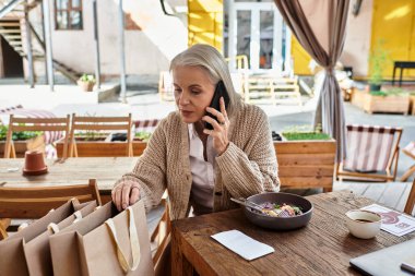 A beautiful mature woman with gray hair engages in a phone conversation while enjoying her meal. clipart