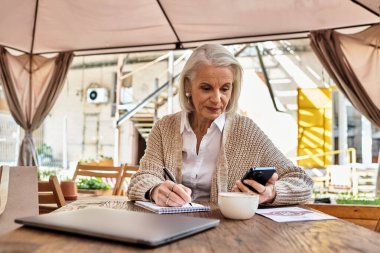 A beautiful gray haired woman enjoys her coffee while writing notes and using her smartphone. clipart