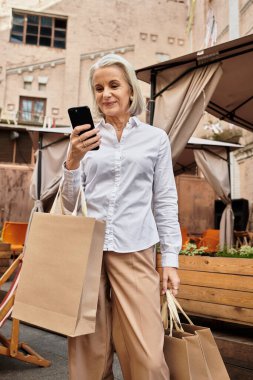 A sophisticated woman checks her phone while carrying shopping bags outdoors in a charming locale. clipart