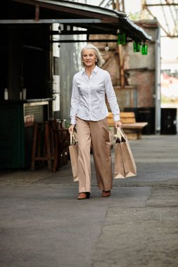 A beautiful mature woman enjoys her stroll, carrying shopping bags down a charming market street. clipart