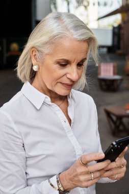 Güzel, olgun bir kadın dışarıda dikilirken akıllı telefonunu dikkatle inceler..