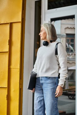 A mature woman with gray hair stands outdoors, carrying a laptop and contemplating her surroundings. clipart