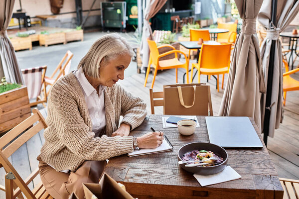 A graceful woman with gray hair deeply engaged in her writing, surrounded by a warm cafe atmosphere.