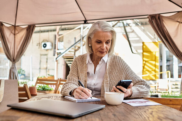 A beautiful gray haired woman enjoys her coffee while writing notes and using her smartphone.