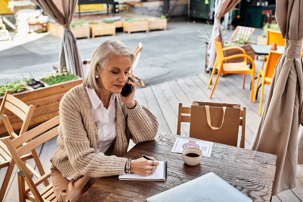 A mature woman is on the phone, taking notes while sipping coffee in a charming cafe.