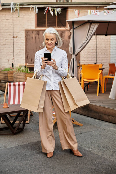Веселая женщина с седыми волосами держит сумки с покупками во время смс в кафе.