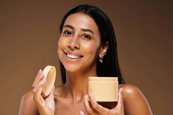 Улыбающаяся брюнетка держит продукт по уходу за кожей в студии, демонстрируя красоту.