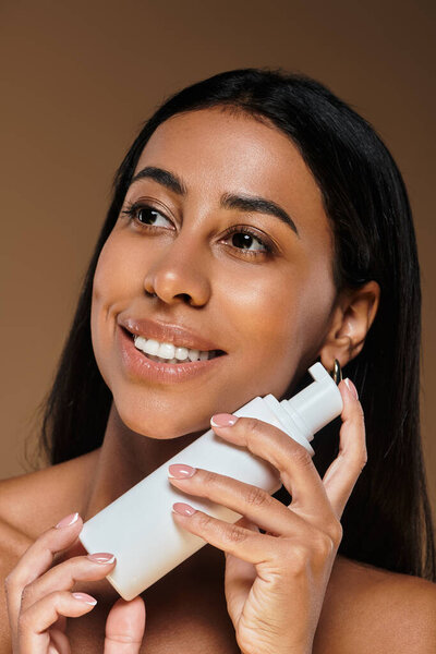 Женщина с брюнетками улыбается, держа продукт по уходу за кожей в студии.