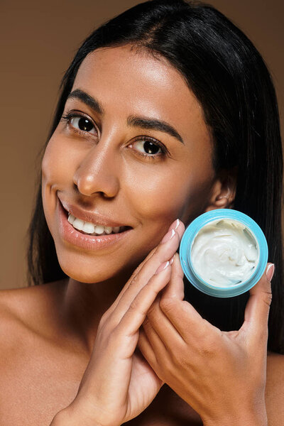 Молодая женщина с брюнетками улыбается, держа продукт по уходу за кожей в студии.