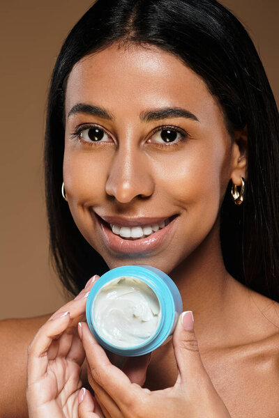 Женщина показывает свою рутину по уходу за кожей, улыбаясь и держа контейнер со сливками в студии.