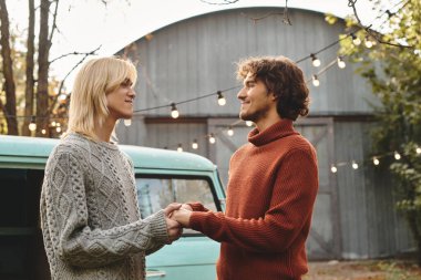 Two young men share a warm gaze while holding hands, surrounded by softly lit autumn ambiance. The backdrop features a vintage green van and trees, creating a romantic atmosphere. clipart