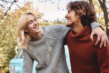 Two young men enjoy a cheerful exchange outdoors surrounded by colorful autumn foliage. Their friendly smiles and relaxed attire reflect a warm connection, highlighting a carefree afternoon together. clipart