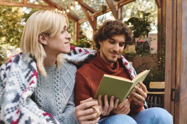 Young loving gay couple wrapped in a blanket share a delightful moment, deeply engaged in a book while surrounded by nature. The cozy garden atmosphere enhances their connection. clipart