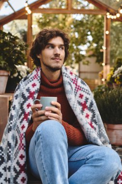 A young and handsome man wrapped in a colorful blanket sits comfortably in a lively greenhouse. clipart