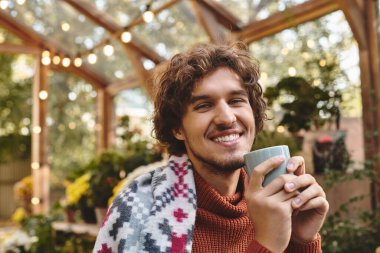 Kıvırcık saçlı genç bir adam, bitkilerle dolu canlı bir serada elinde fincan tutarken sıcak bir şekilde gülümsüyor. Güneş ışığı cam panellerden süzülüp etrafında neşeli bir atmosfer yaratıyor..