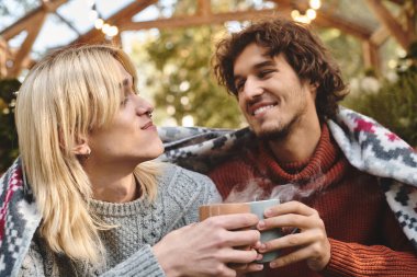 In a serene outdoor setting, two young and handsome men enjoy each others company while sharing warm drinks. The cozy atmosphere, enhanced by a blanket, reflects their close bond and comfort. clipart