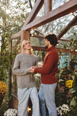 In a charming greenhouse filled with vibrant flowers, a young couple shares a joyful connection. Dressed in stylish sweaters, they hold hands, exuding warmth and affection in the serene atmosphere. clipart