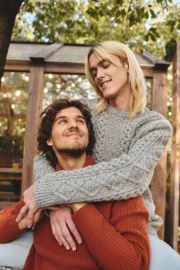 Huzurlu bir bahçe ortamında, genç bir çift, şık kazaklarıyla mutluluk saçıyor. Yumuşak sonbahar ışığı, birbirlerine eşlik ederken neşeli ifadelerini vurgular..