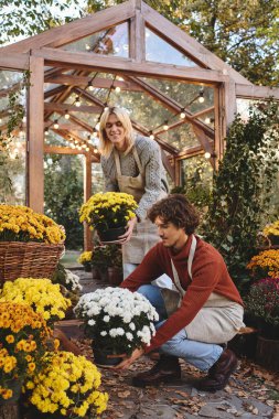 Young loving gay couple joyfully arrange colorful potted flowers inside a cozy greenhouse. Surrounded by lush greenery and warm lights, they share a moment of creativity and love for nature. clipart