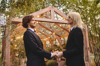 In a lush garden setting, two young men dressed in elegant suits exchange heartfelt vows while holding hands. Surrounded by greenery and soft lights, the atmosphere radiates romance and joy. clipart